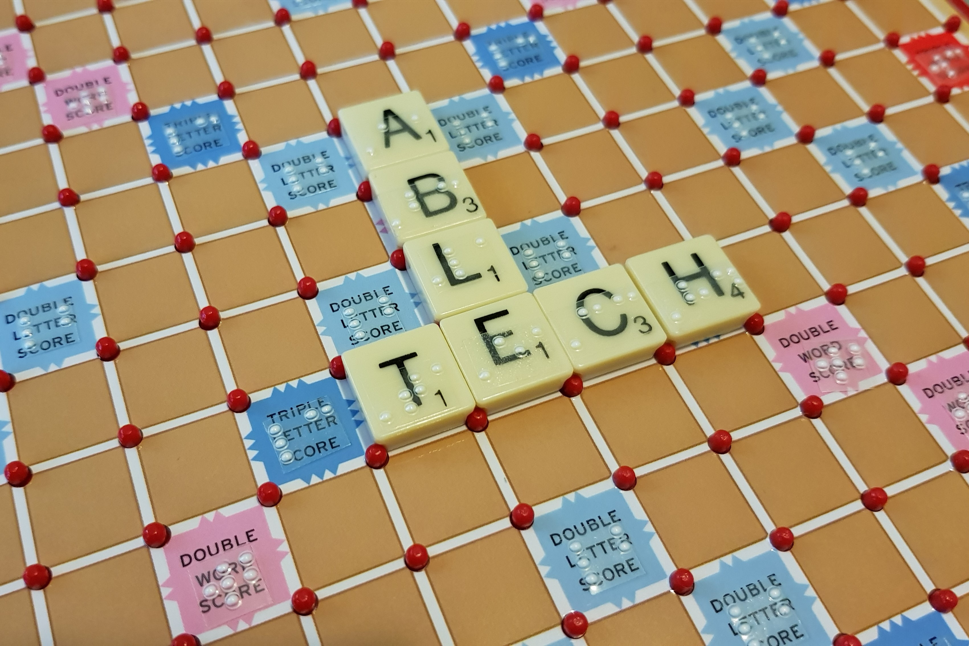 A Scrabble board and letter tiles labelled with braille. The letter tiles placed on the board have the word “TECH” formed across the middle of the board, with the word “ABLE” formed on top of the letter “E”.