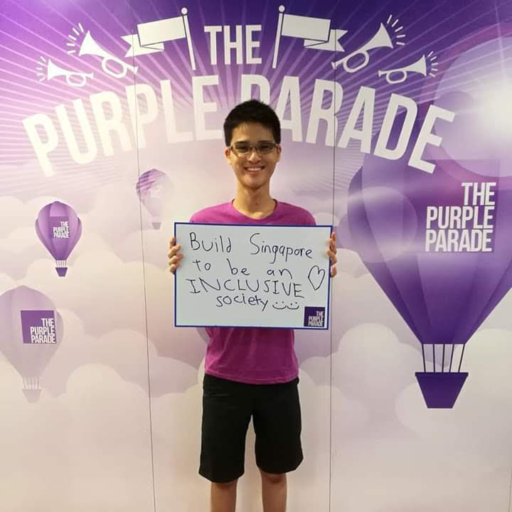 The photo shows Wesley in a purple t-shirt holding up a sign that says “Build Singapore to be an INCLUSIVE society  ” in front of a backdrop with “The Purple Parade” written several times on it.